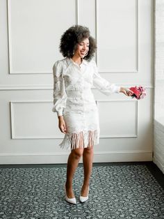 a woman in a white dress is smiling and holding a pink flower while standing next to a wall