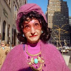 an old woman wearing glasses and a purple sweater in front of tall buildings with people walking down the street