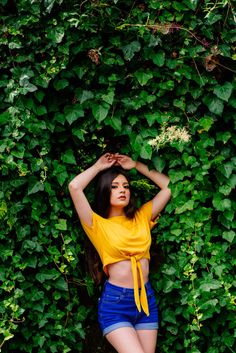 a woman in yellow shirt and blue shorts standing next to green plants with her hands on her head