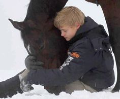 a young boy kneeling down next to a horse in the snow with it's head on his hand