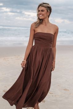 a woman standing on top of a sandy beach next to the ocean wearing a brown dress