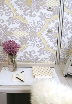 a vase with flowers on a desk in front of a wallpapered background and writing utensils