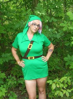 a woman dressed in a green outfit standing next to some bushes and trees with her hands on her hips