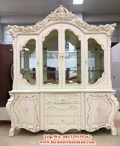 a white china cabinet with glass doors and ornate carvings on the front, sitting in a room