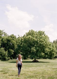 a woman walking through a field with trees in the background