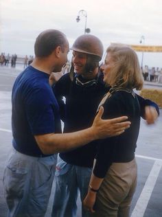 two men and a woman standing in a parking lot talking to each other with their arms around one another