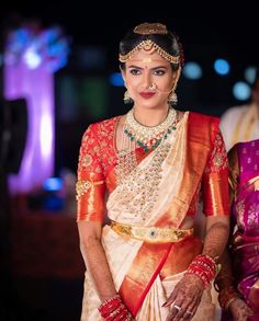 a woman in a red and gold sari with jewelry on her head, standing next to another woman