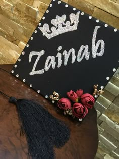 a black sign that says zainab with flowers on it and a tassel