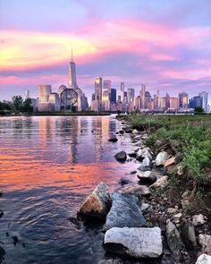 the city skyline is reflected in the water at sunset, with rocks and boulders on the shore