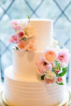 a three tiered white cake with pink and white flowers on the top, sitting in front of a window