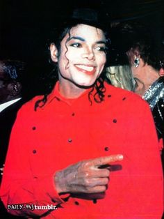 michael jackson in red shirt and black hat with his hand on his hip, smiling at the camera