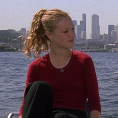 a woman sitting on top of a boat in the water with a city behind her