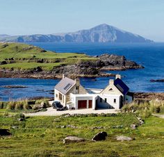 a white house sitting on top of a lush green hillside next to the ocean with mountains in the background