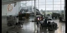 two men standing in front of an airplane inside of a hanger filled with vehicles