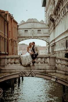 a bride and groom sitting on a bridge over a river in an old european city