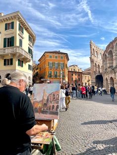 an older man is painting in front of some old buildings and people are walking around