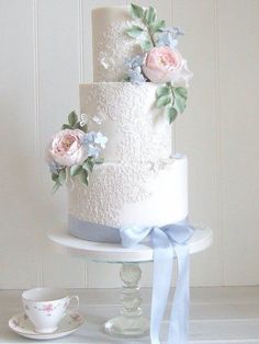 a white wedding cake with pink roses and blue ribbon on the top is surrounded by teacups