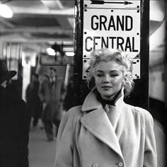 a black and white photo of a woman standing in front of a grand central sign