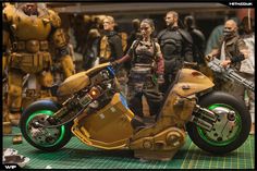 a group of action figures standing around a toy motorcycle with people in the back ground