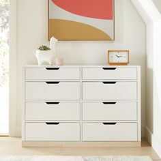 a white dresser with black drawers and a clock on the wall next to an art piece