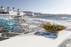 a bowl of fruit sitting on top of a white table next to the ocean with blue chairs