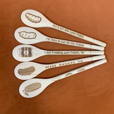 four wooden spoons with different designs on them sitting on a brown surface, one says i'm here to eat all the pickles