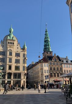 people are walking around in an old town square with tall buildings and green spires