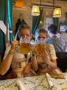 two women sitting at a table with beer glasses in front of their faces and smiling