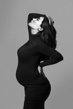 a pregnant woman in a black dress poses with her hands on her head and arms behind her back