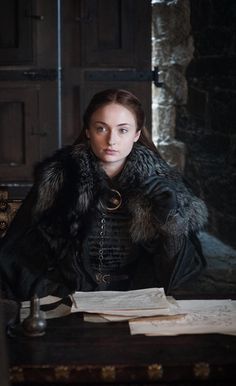 a woman sitting at a table in a room with papers on it and wearing a fur collar