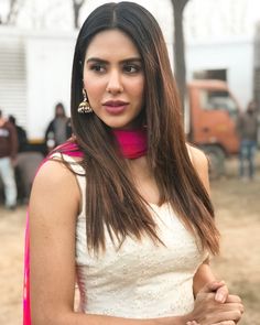 a woman with long hair wearing a white dress and pink scarf around her neck is looking at the camera