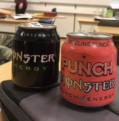 two cans of monster energy and punch energy sit on a backpack in an office cubicle