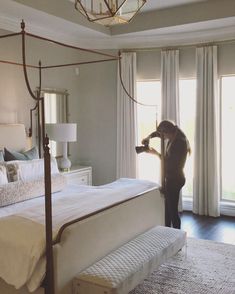 a woman standing in front of a bed with white sheets and pillows, looking out the window