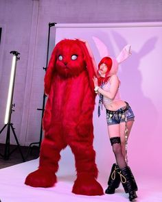 a woman standing next to a giant red furry animal in front of a pink backdrop