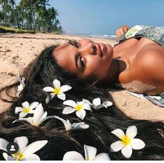 a beautiful woman laying on top of a sandy beach next to the ocean with white flowers in her hair