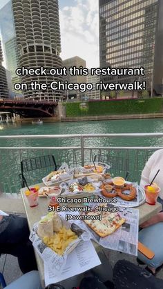 two people sitting at a table with food and drinks on it in front of the chicago river