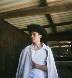 a young man wearing a cowboy hat and white shirt standing next to a wooden wall