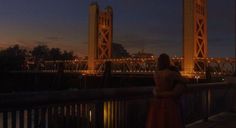 a woman in a long dress standing on a bridge looking at the water and buildings