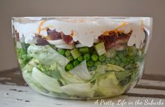 a salad in a glass bowl with peas, bacon, lettuce and cheese