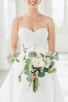 a bride holding a bouquet of white flowers and greenery in her wedding dress smiling at the camera