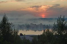 the sun is setting over a lake surrounded by trees and fog in the distance, with birds flying overhead