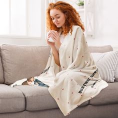 a woman wrapped in a blanket sitting on a couch drinking from a cup while holding a mug