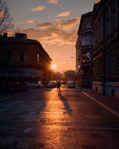 the sun is setting over a city street with cars parked on it and people walking down the sidewalk