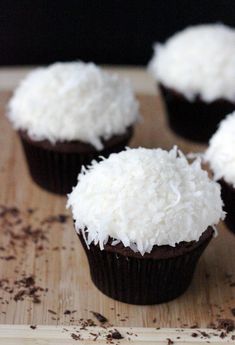 four cupcakes topped with white frosting and coconut on a wooden table top