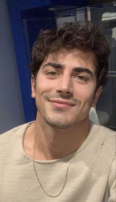 a man with curly hair smiling at the camera while wearing a tan sweater and gold necklace