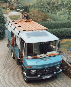 a man sitting on top of a blue van parked next to a lush green field