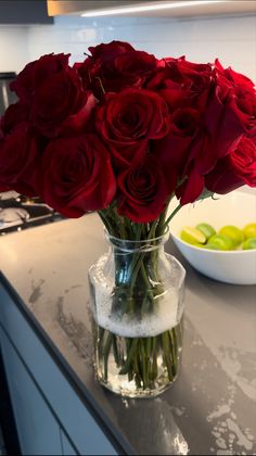 a vase filled with red roses sitting on top of a counter next to a bowl of fruit