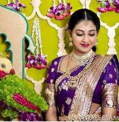 a woman in a purple and gold sari holding a green plant with flowers on it