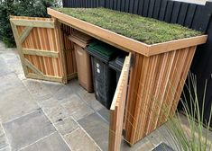 a wooden shed with grass growing on the roof and trash cans in it's bins