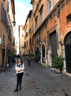 a woman standing in the middle of an alleyway with buildings on both sides and people walking around